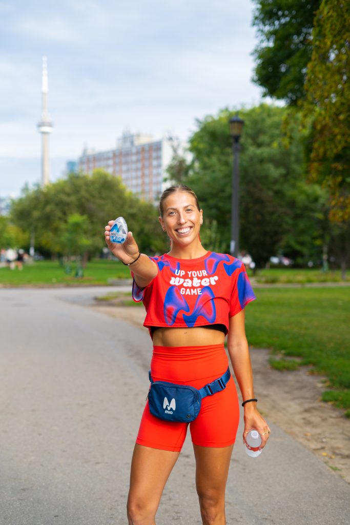 FX Productions Canada A woman in a red athletic outfit holds out a water bottle and smiles while standing on a park path; trees and buildings are visible in the background. Toronto Video Production Company