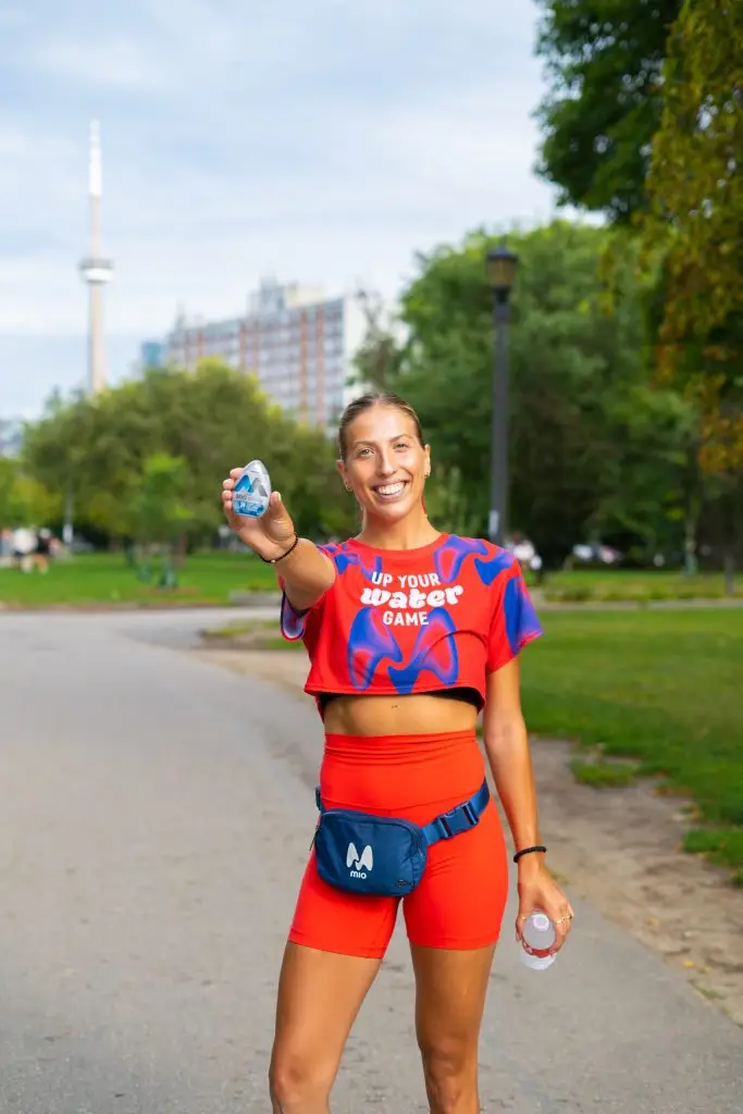FX Productions Canada A woman in a red athletic outfit holds out a water bottle and smiles while standing on a park path; trees and buildings are visible in the background. Toronto Video Production Company