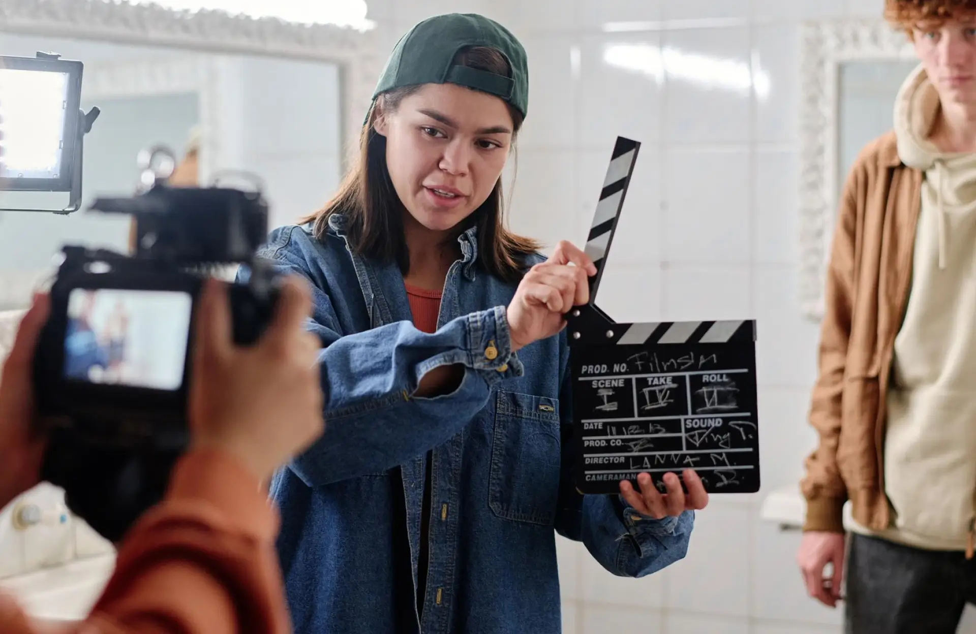 FX Productions Canada Person in a denim jacket and backwards cap holds a clapperboard in front of a camera, preparing to film a scene in a brightly lit room. Toronto Video Production Company