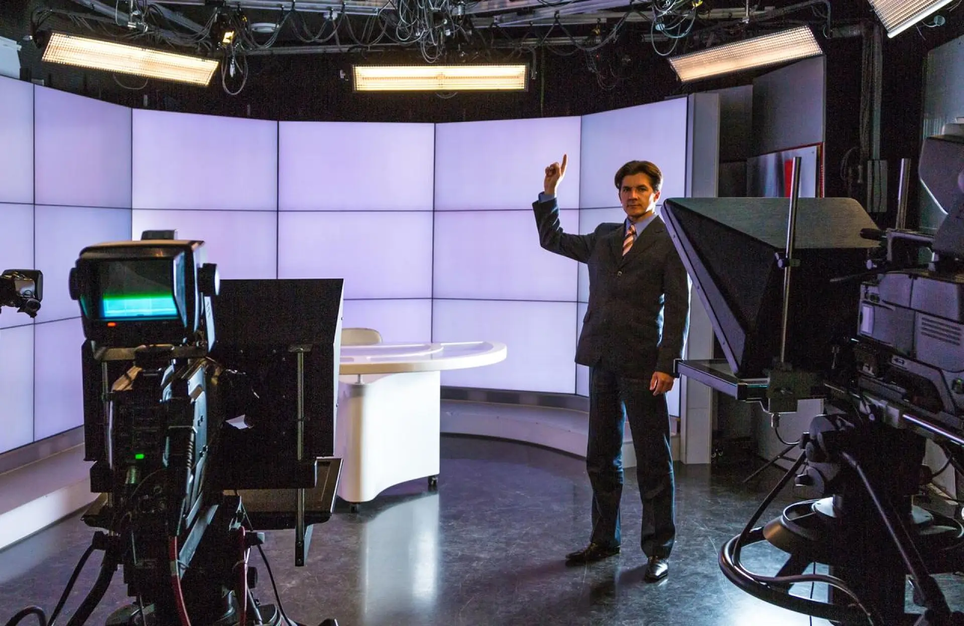 FX Productions Canada A person in a suit stands in a television studio, raising one hand, surrounded by cameras and studio lighting, highlighting the professionalism of video production services in front of a lit panel backdrop. Toronto Video Production Company
