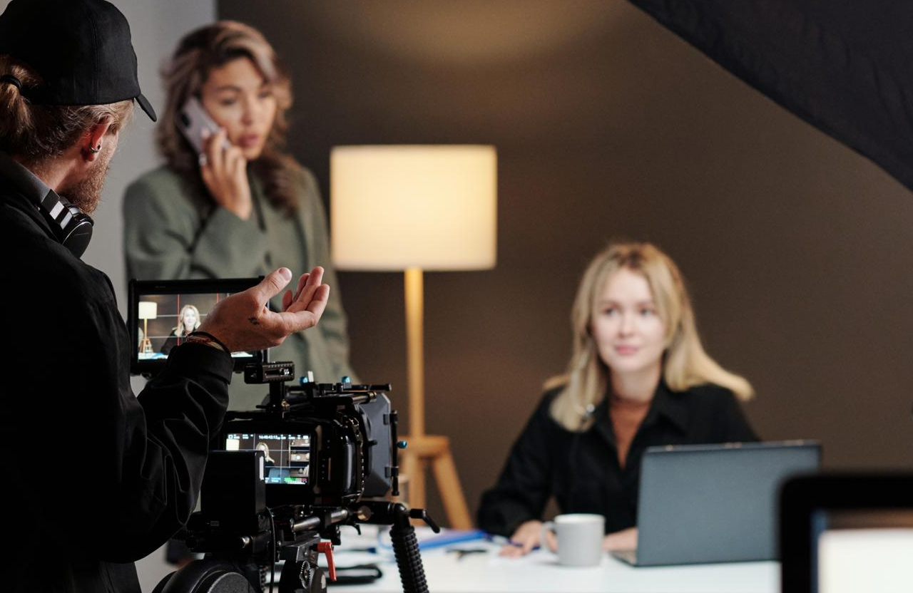 FX Productions Canada A person films two women, one sitting at a desk with a laptop and coffee cup, the other standing and talking on the phone, with a lamp in the background—perfect for cinematic storytelling in corporate video production service. Toronto Video Production Company