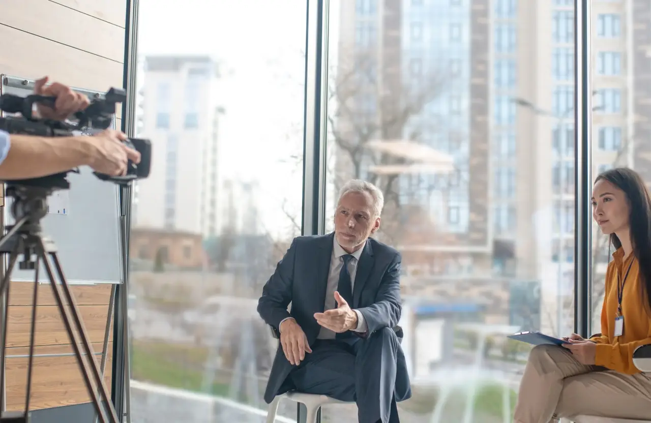 FX Productions Canada An older man in a suit is interviewed while a woman takes notes and a camera crew films them in a modern office with large windows, showcasing cinematic storytelling through professional video production services. Toronto Video Production Company