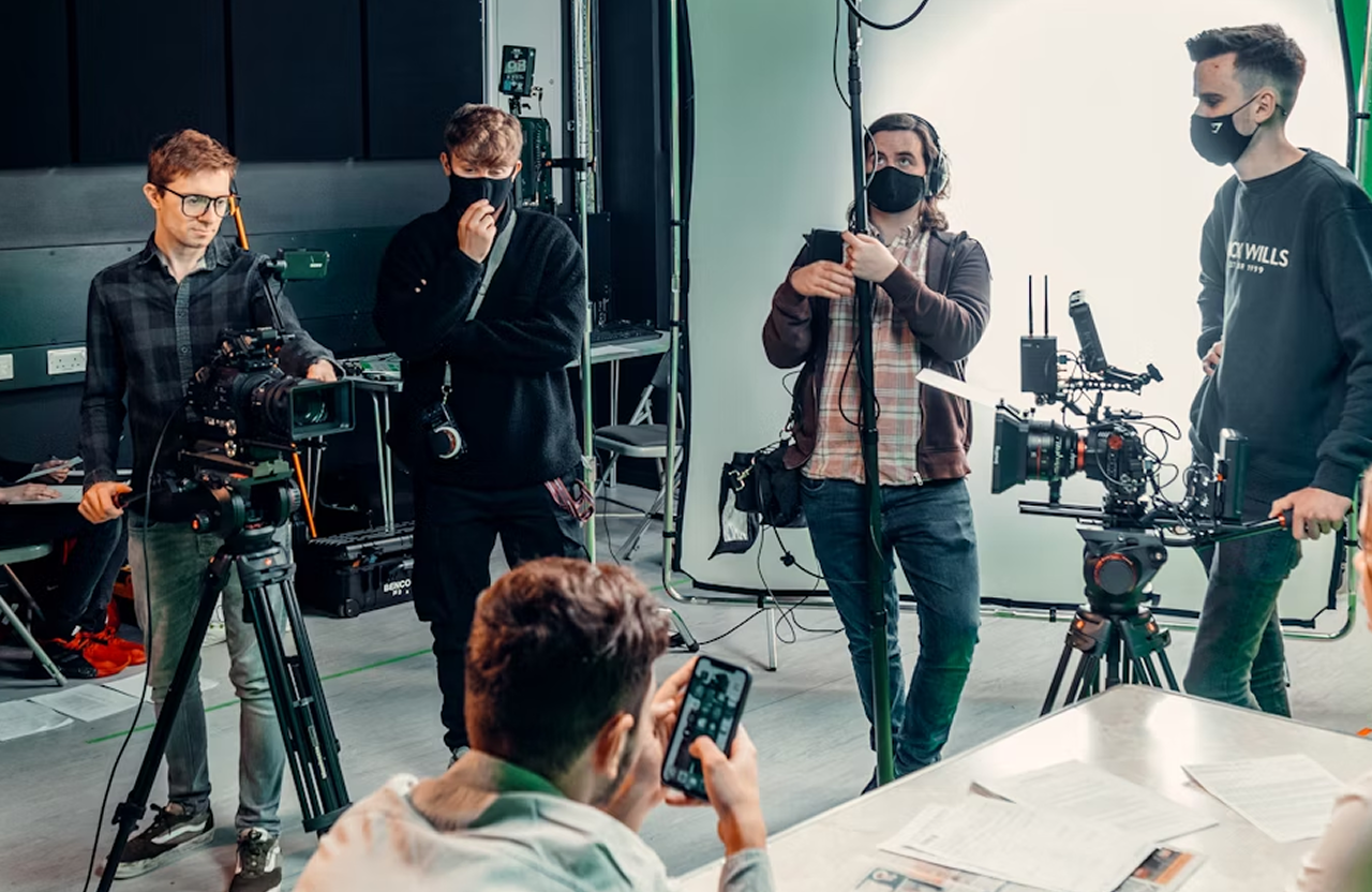 FX Productions Canada Four people from a creative video agency with film equipment and face masks stand around a seated person using a smartphone, in a studio setting with lights and cameras visible, showcasing cinematic storytelling in action. Toronto Video Production Company