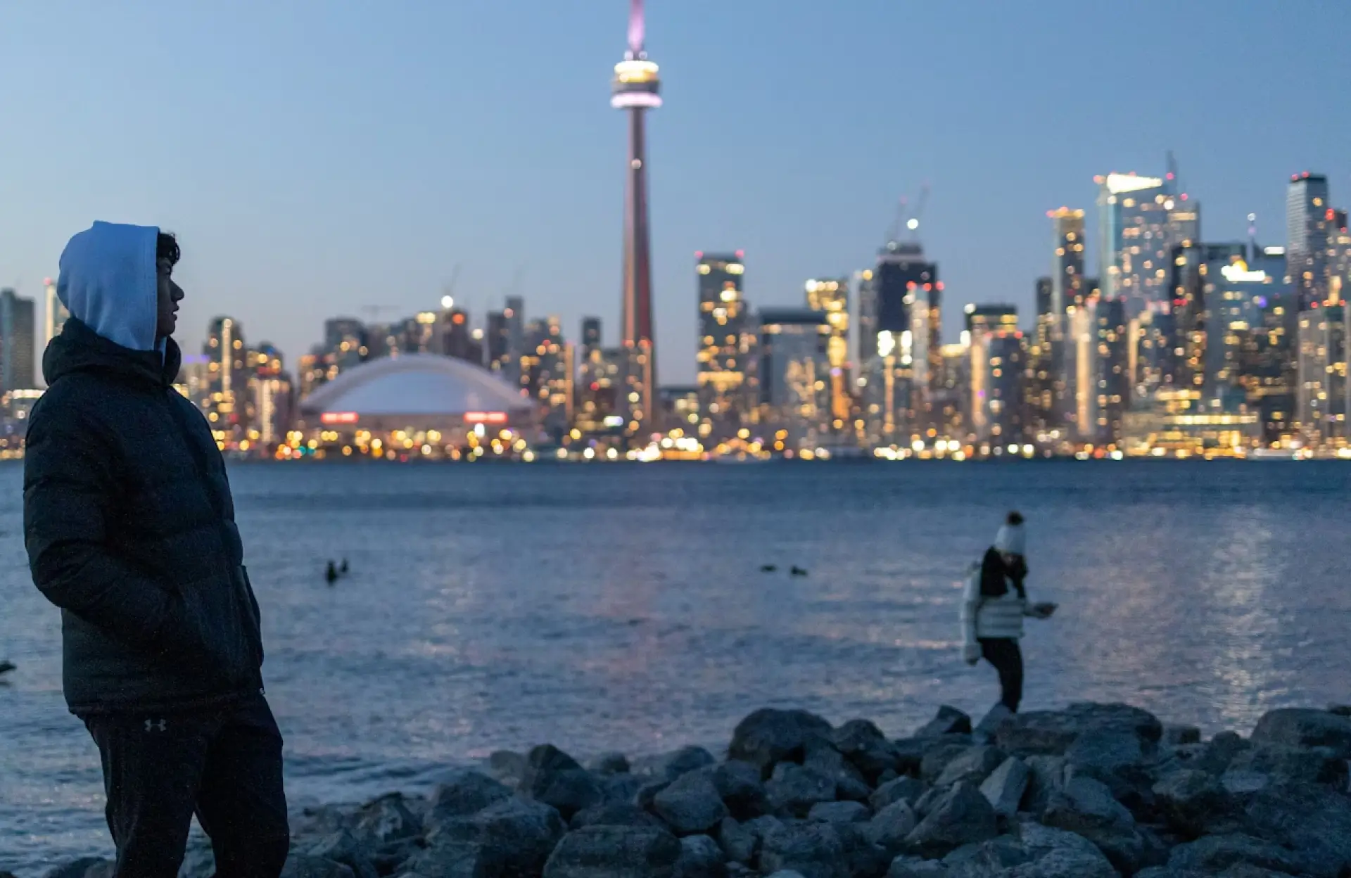 FX Productions Canada Two people stand on a rocky shore at dusk with Toronto skyline, CN Tower, and city lights in the background across the water. Toronto Video Production Company