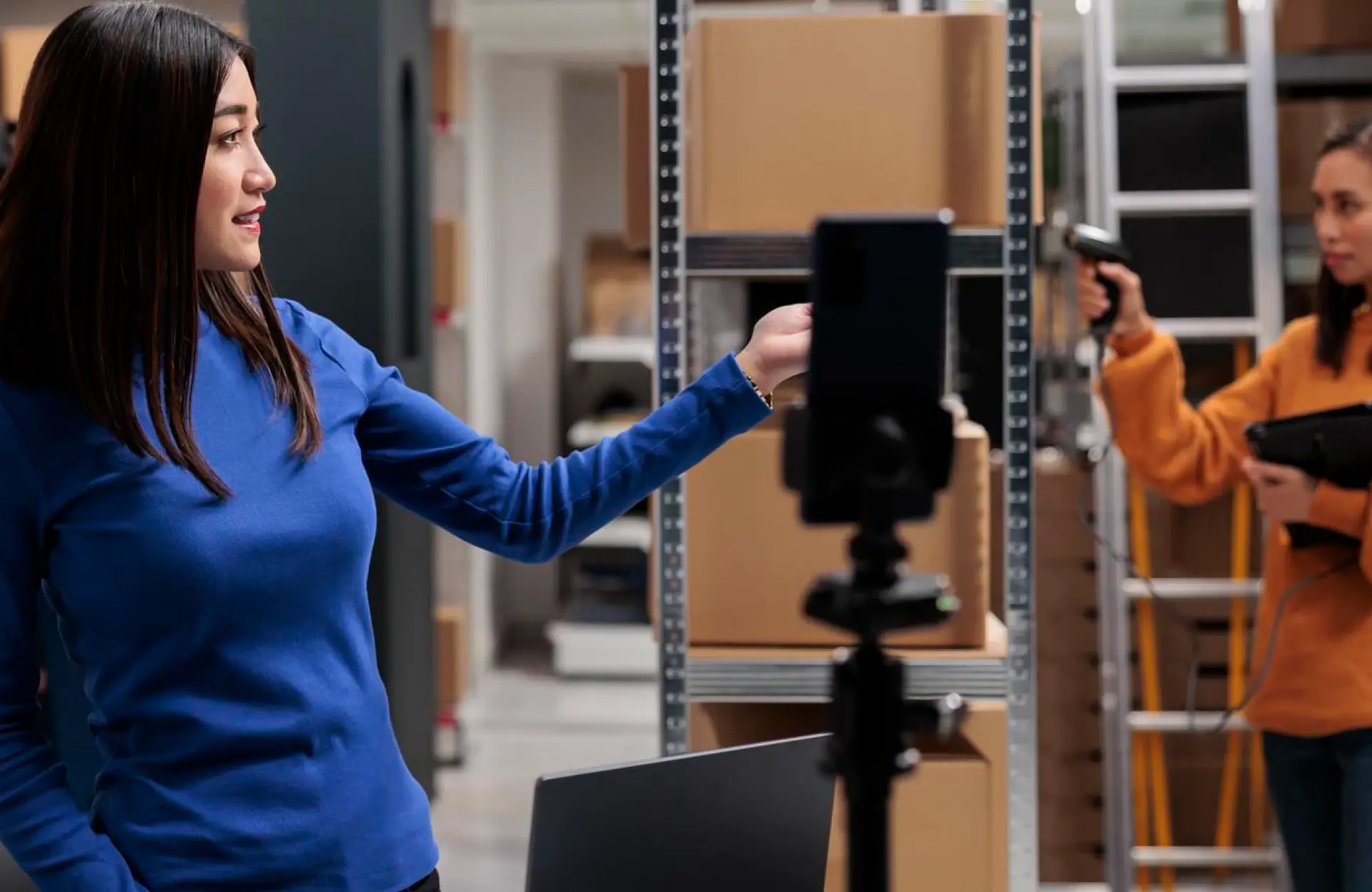 FX Productions Canada Two women are working in a storage area; one stands near a camera and gestures, while the other uses a handheld scanner by metal shelves with cardboard boxes. Toronto Video Production Company