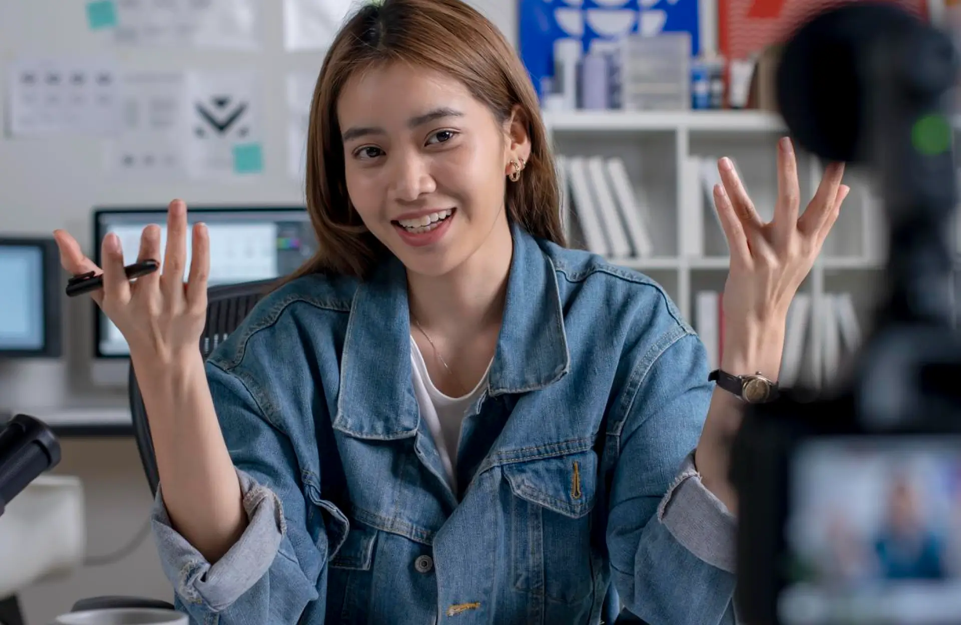 FX Productions Canada A woman in a denim jacket speaks while gesturing with her hands, sitting at a desk with a pen, in front of a camera and computer monitors. Toronto Video Production Company