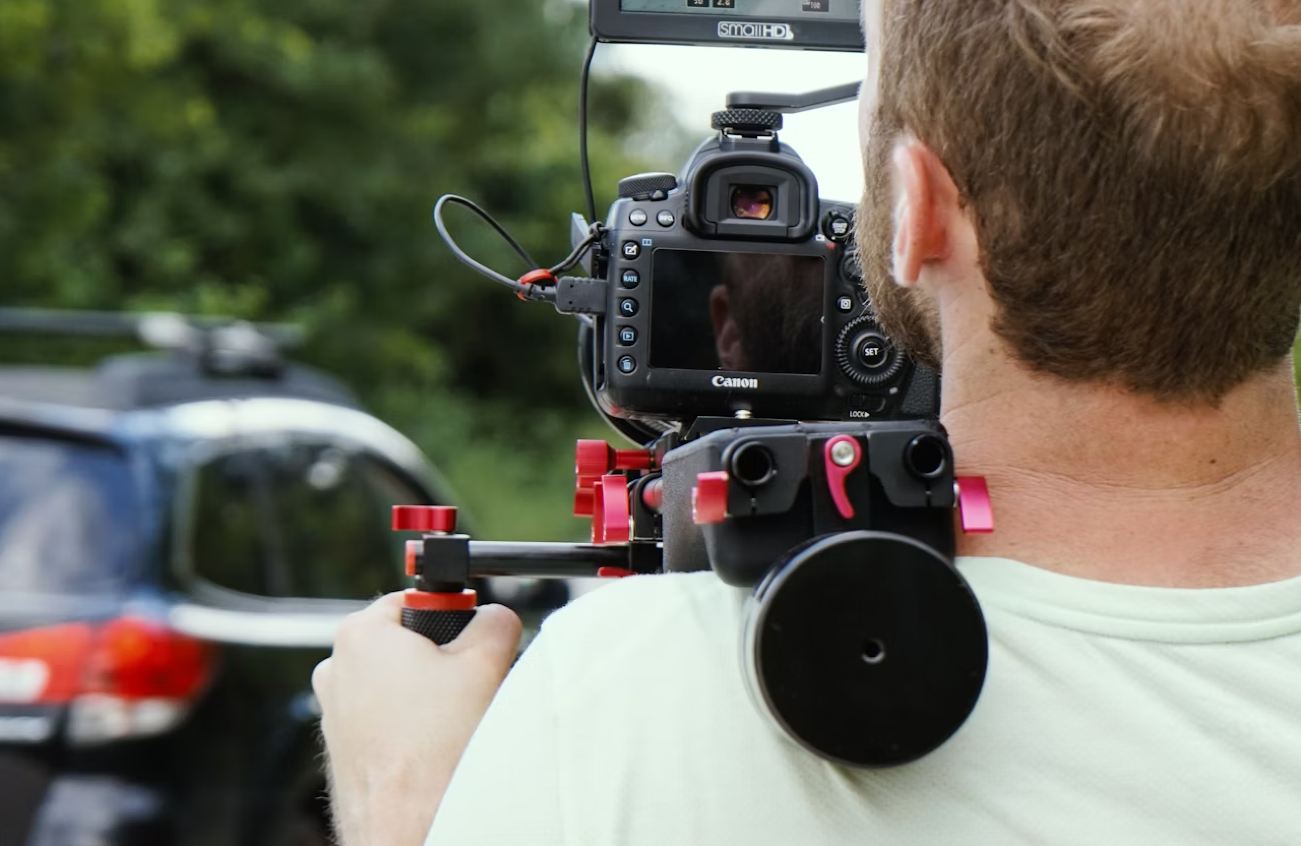 FX Productions Canada A person operates a Canon DSLR camera mounted on a shoulder rig, filming a dark-colored car outdoors near greenery for a post production company. Toronto Video Production Company