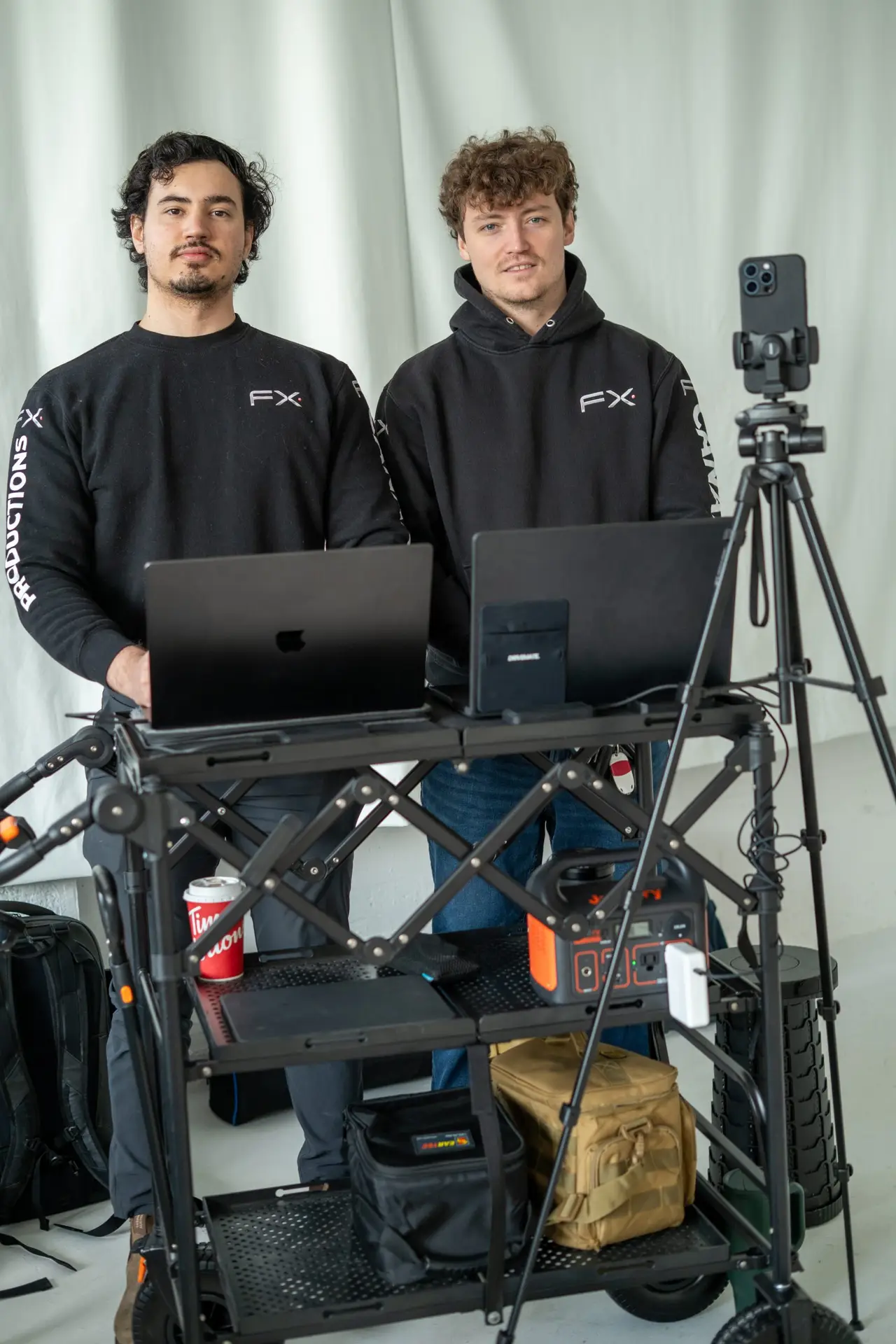 FX Productions Canada Two men stand behind a rolling cart with laptops and gear; a smartphone is mounted on a tripod in front of them against a plain backdrop. Toronto Video Production Company