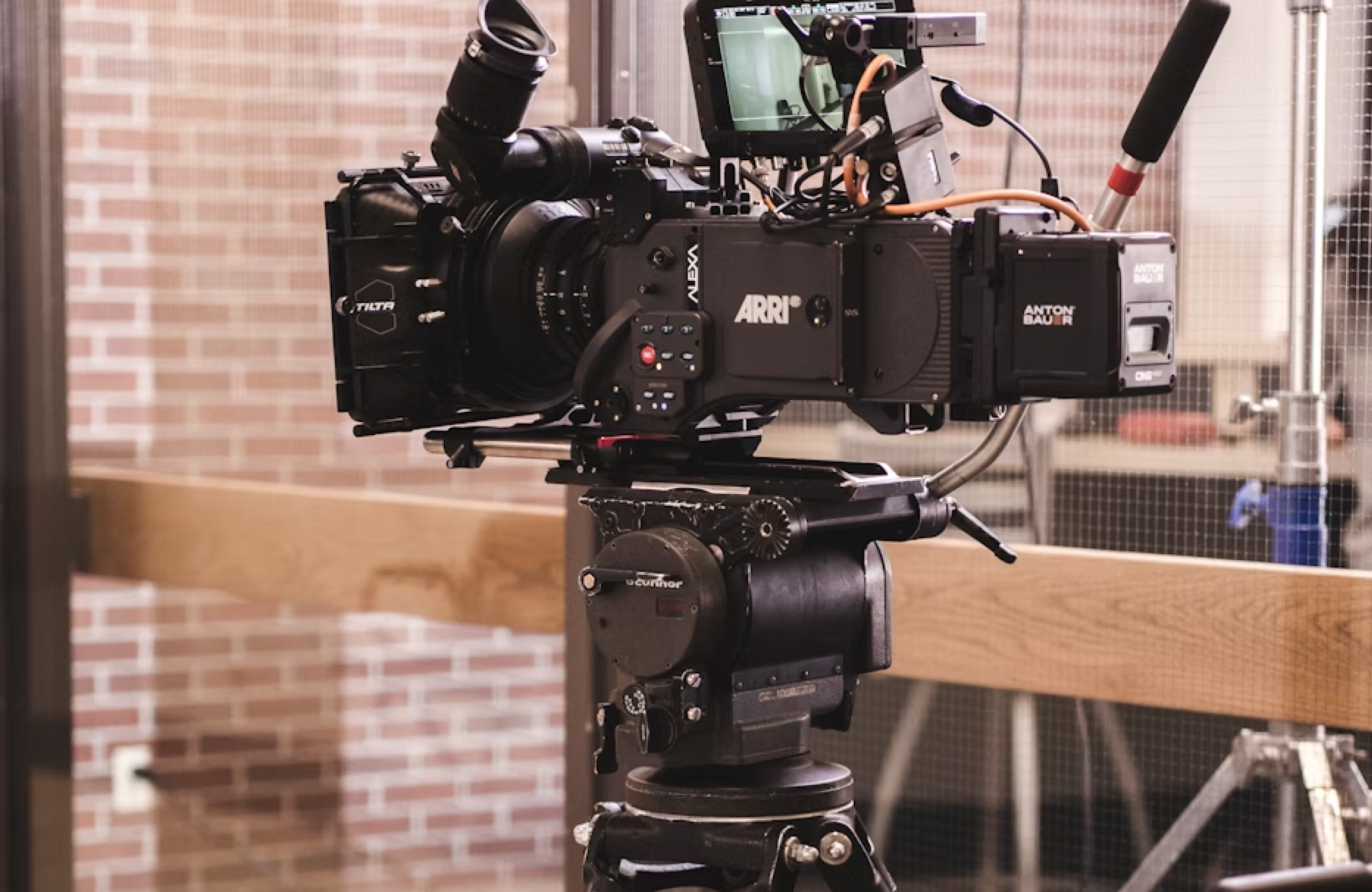 FX Productions Canada A professional video camera setup with various attachments is mounted on a tripod indoors, with a brick wall and glass door in the background. Toronto Video Production Company