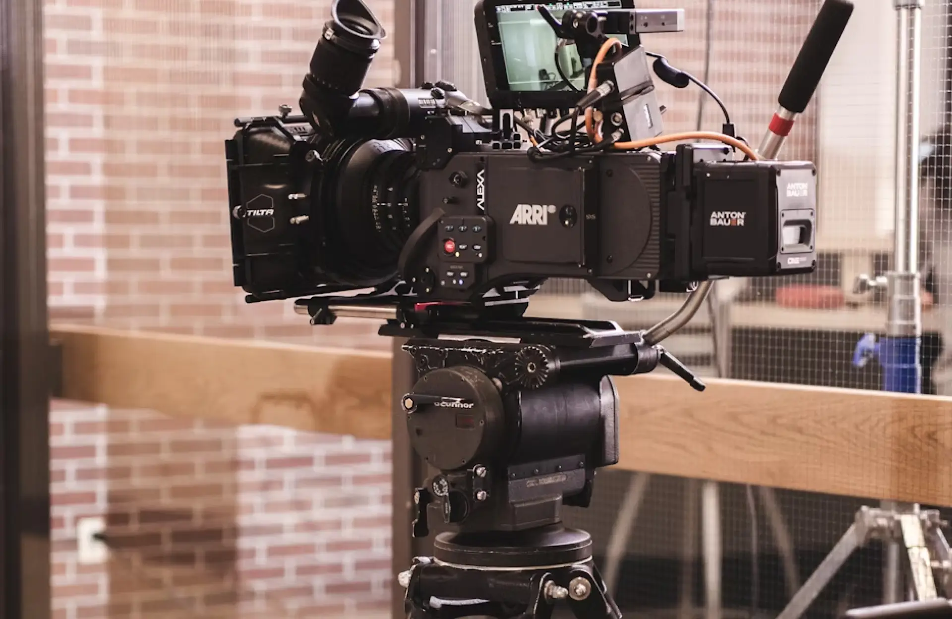 FX Productions Canada A professional video camera setup with various attachments is mounted on a tripod indoors, with a brick wall and glass door in the background. Toronto Video Production Company
