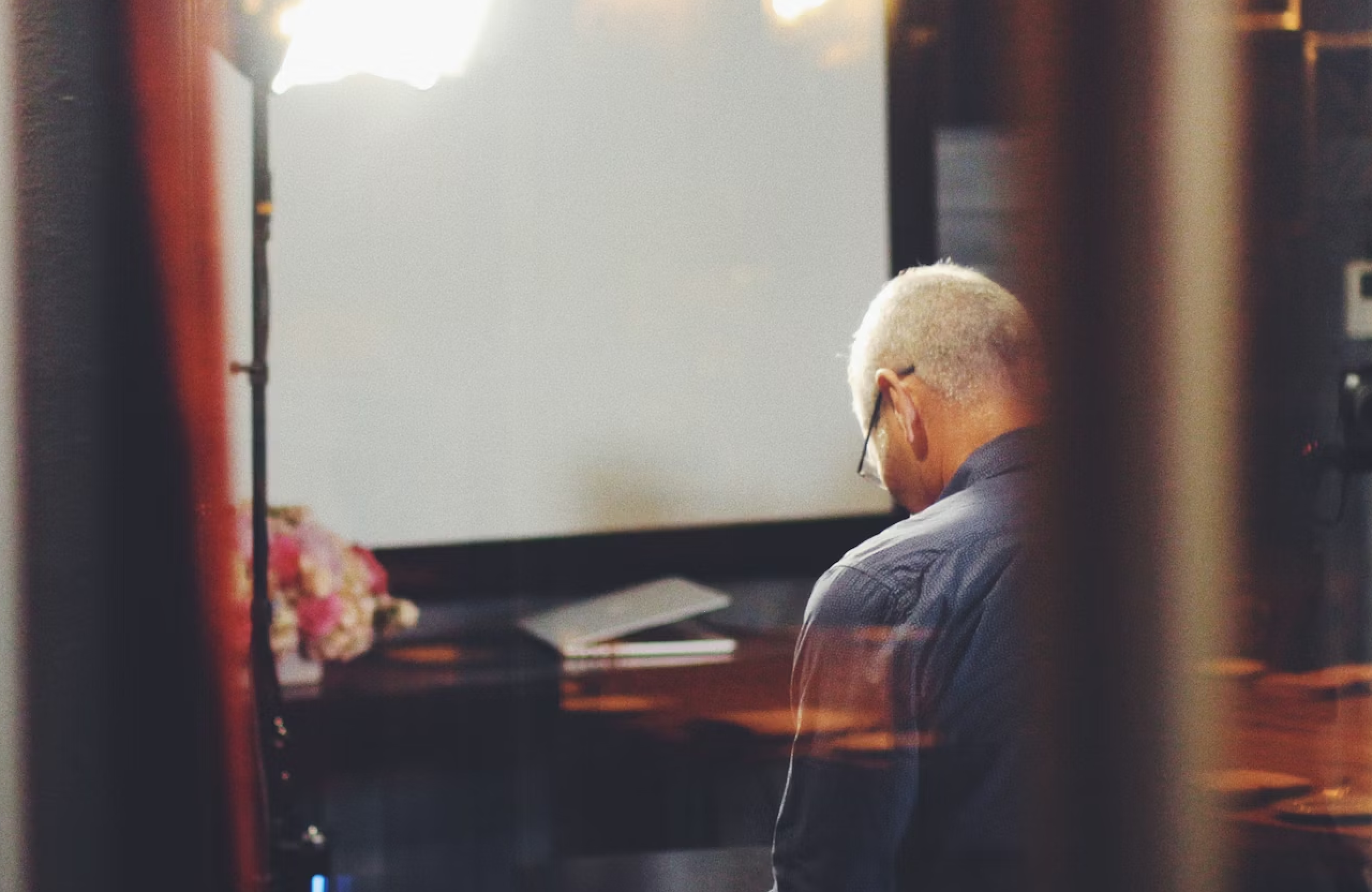FX Productions Canada An older man with gray hair sits alone at a table in a dimly lit room, facing away from the camera. A bright light source and a laptop are visible in the background. Toronto Video Production Company