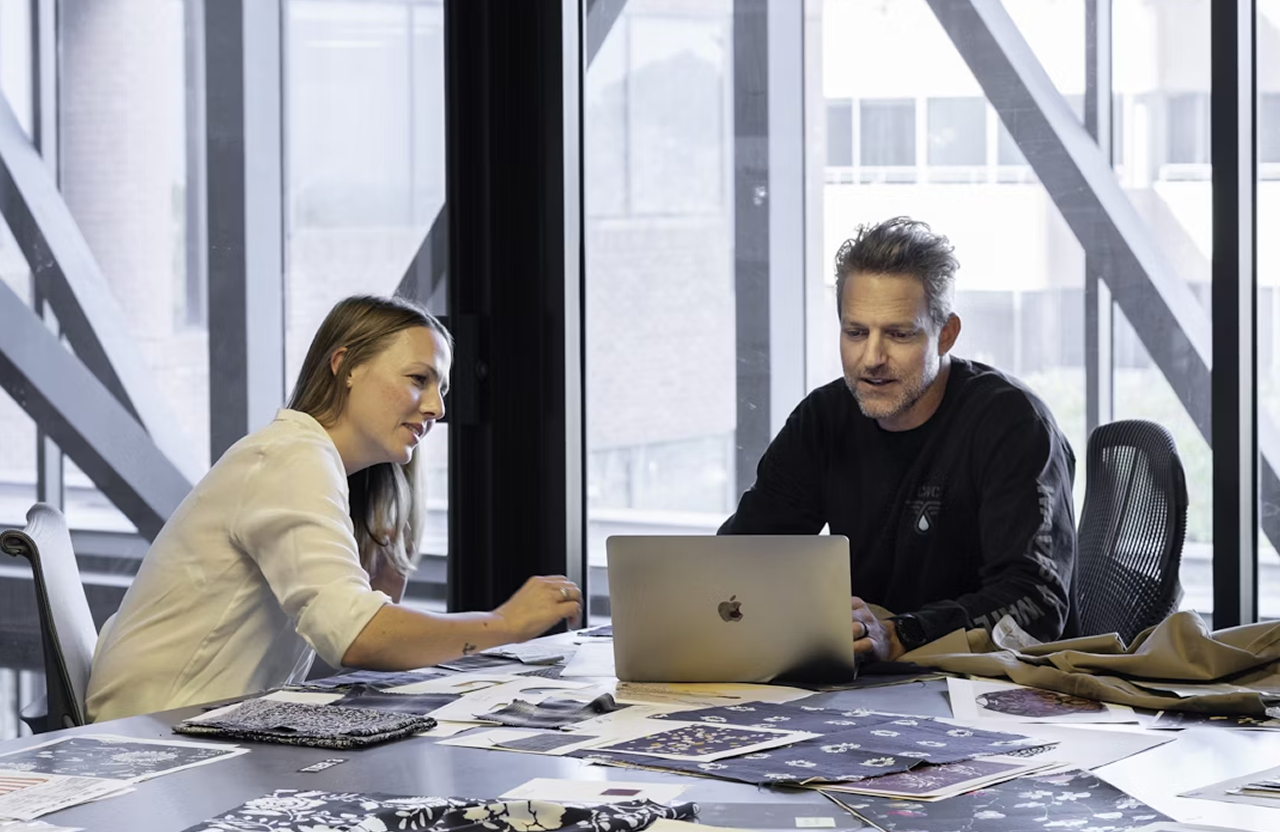 FX Productions Canada Two people sit at a table covered with papers and fabric samples, looking at a laptop together in a modern office with large windows. Toronto Video Production Company