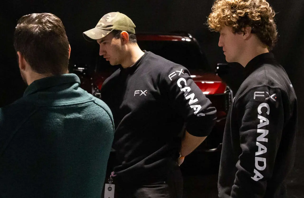 FX Productions Canada Three men stand indoors near a red vehicle. Two wear black FX Canada sweatshirts, and one wears a green cap. The background is dimly lit. Toronto Video Production Company