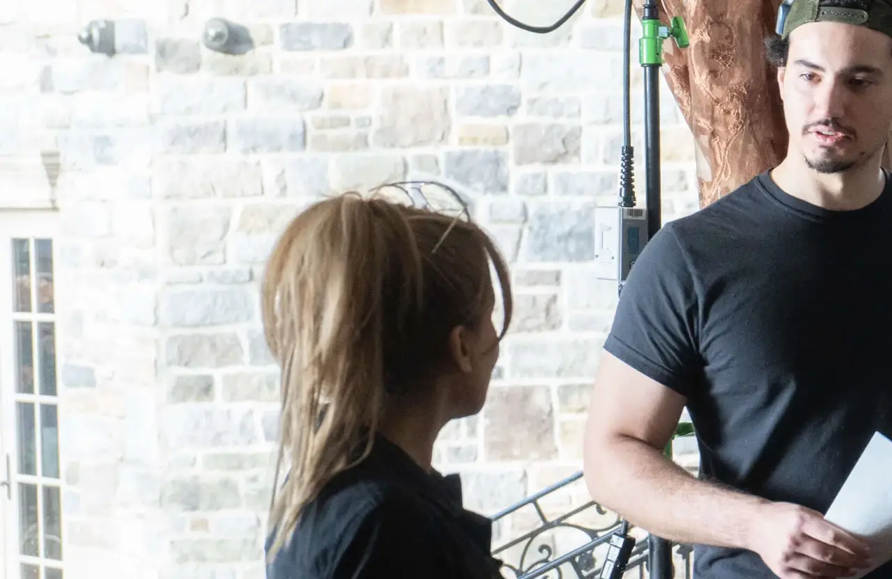 FX Productions Canada Two people stand indoors near a stone wall—one with a ponytail facing away, the other holding a paper—collaborating on a project for a full service video production in Toronto. Toronto Video Production Company