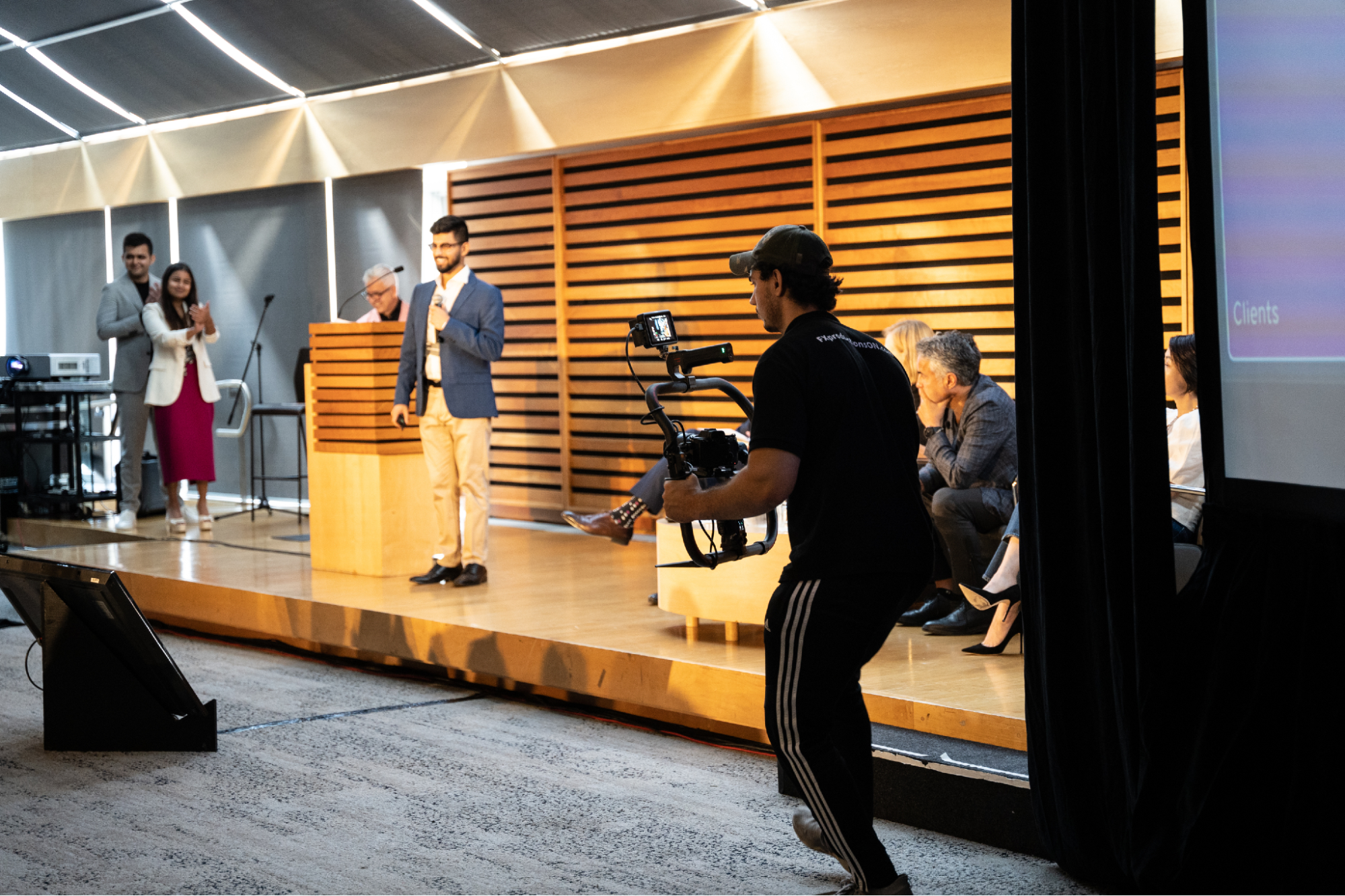 FX Productions Canada A cameraman from a production partner in Toronto films a group on stage during a presentation in a modern conference room, capturing key moments for investor relations video production in Toronto. Some speak on stage while others sit and watch. Toronto Video Production Company