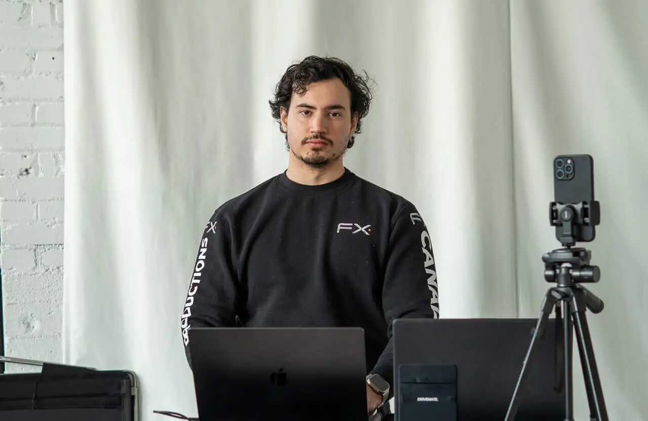FX Productions Canada Man with dark hair and a black sweatshirt sits at a desk with two laptops and a smartphone on a tripod, against a light curtain background, working on automotive video production Toronto projects. Toronto Video Production Company