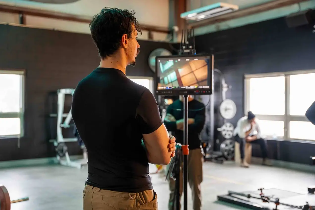 FX Productions Canada A man stands with arms crossed, looking at a monitor in a gym setting, while another person sits by the windows in the background. Toronto Video Production Company