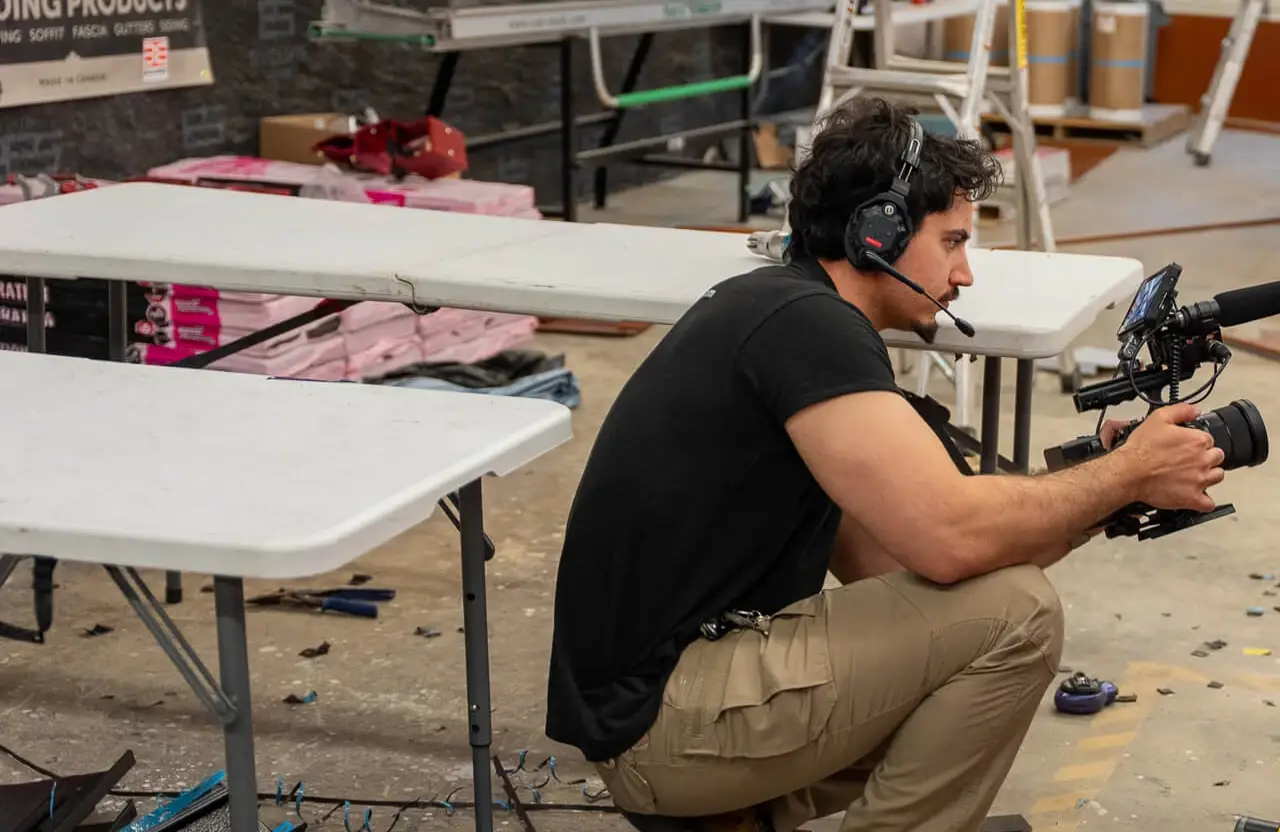 FX Productions Canada A man wearing a headset crouches next to folding tables, holding a professional video camera in a workshop or warehouse setting. Toronto Video Production Company
