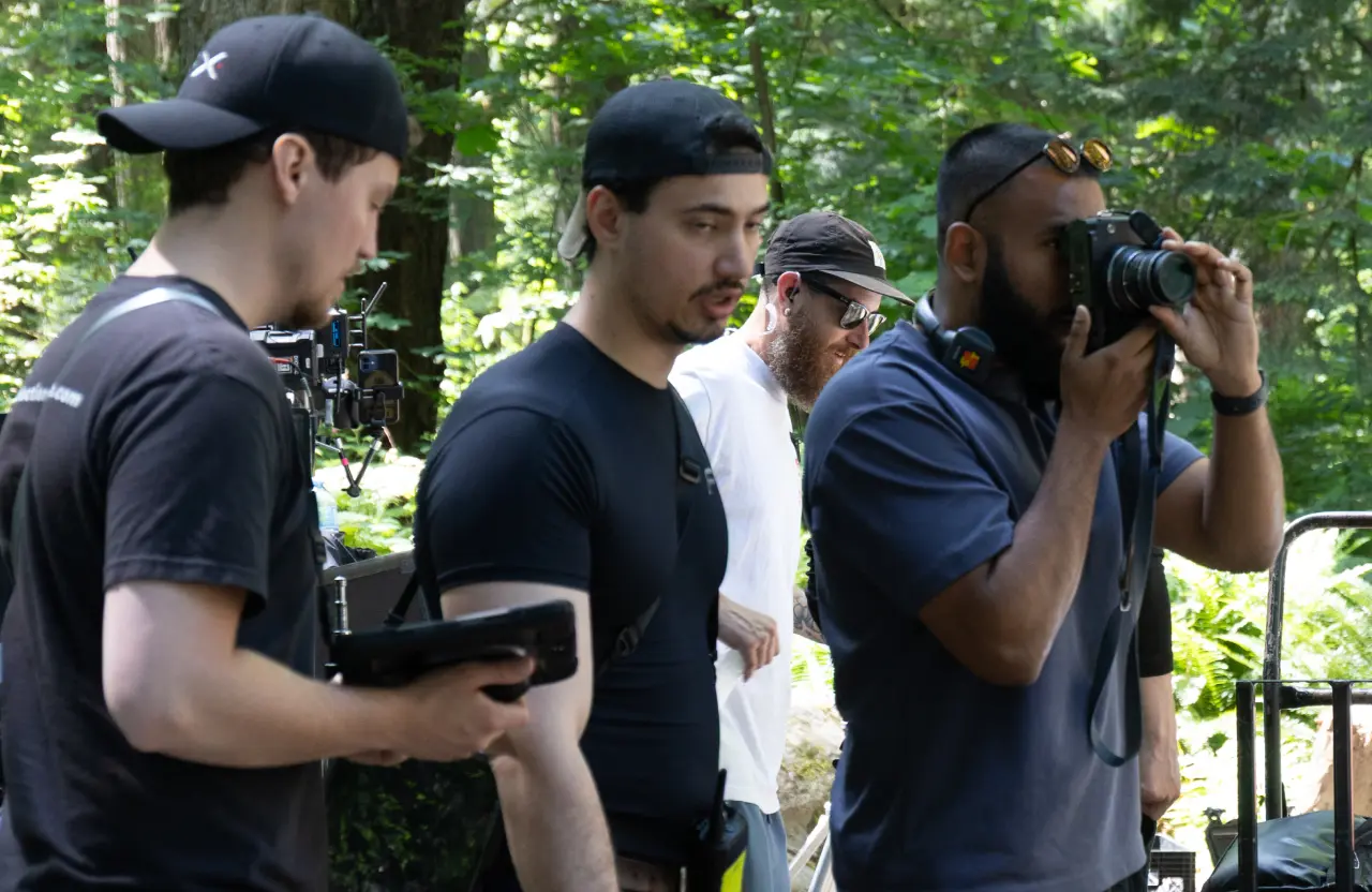 FX Productions Canada Four men with film equipment stand outdoors; one looks through a camera while others watch, surrounded by trees and natural light—a behind-the-scenes look at video production pricing models in Toronto. Toronto Video Production Company