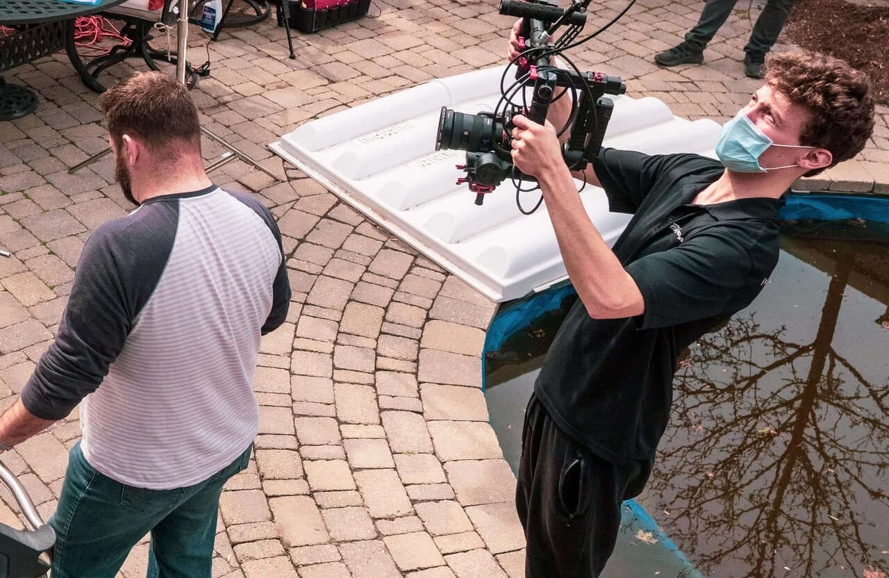 FX Productions Canada A person wearing a mask films with a camera rig outdoors near a small pond, while another person stands nearby facing away—capturing the essential steps to build a video production roadmap. Toronto Video Production Company