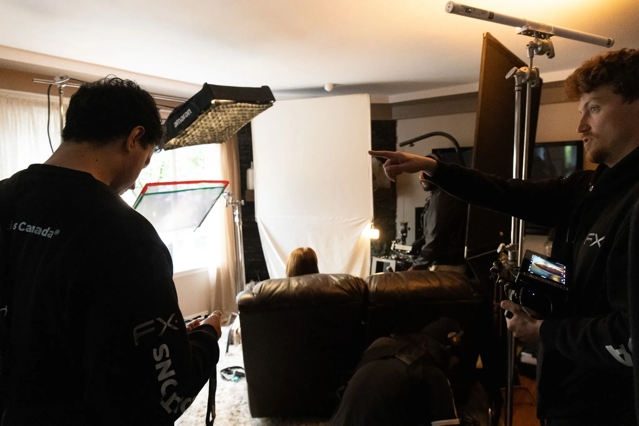 FX Productions Canada Two crew members from a commercial production company BC set up filming equipment in a living room, with one person seated facing a bright white backdrop between two couches. Toronto Video Production Company