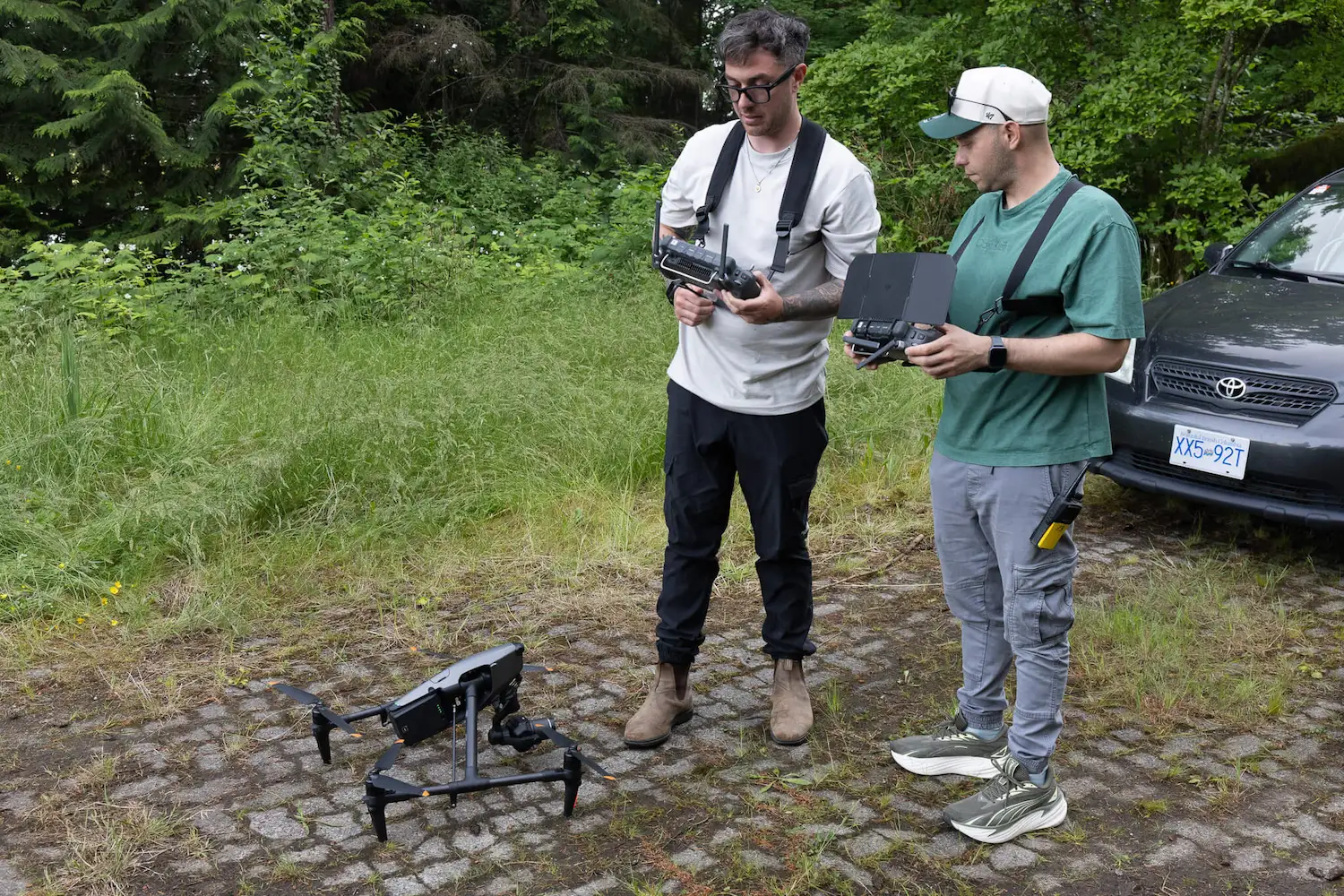 FX Productions Canada Two people stand outdoors on a stone path, operating a large drone with remote controllers—demonstrating professional drone services in Vancouver. Trees and a parked car are visible in the background. Toronto Video Production Company