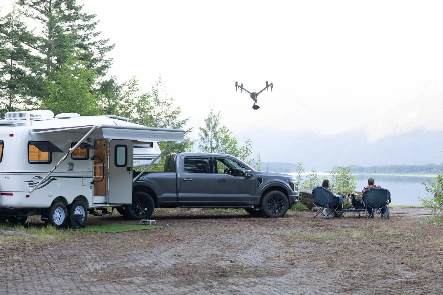 FX Productions Canada A pickup truck and camper are parked by a lake. Two people sit in chairs facing the water, while a drone filming Vancouver captures the tranquil scene from above. Trees and mountains are visible in the background. Toronto Video Production Company